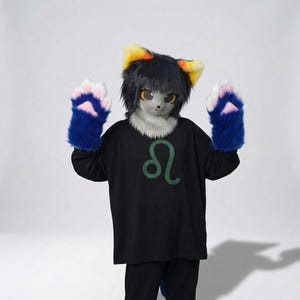 Può includere: Una persona in un costume fursuit simile a un gatto. Il costume presenta una testa grigia con capelli neri e orecchie gialle e arancioni. La persona indossa una maglietta nera con un simbolo verde e guanti blu a forma di zampa.