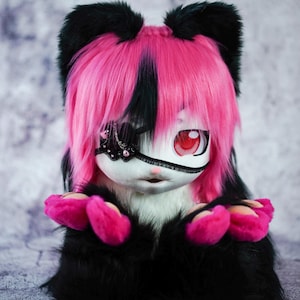May include: A plush toy with black and pink fur, wearing a pink wig with a black streak. The toy has a white face with a black eye patch and one red eye. The toy is sitting with its paws in front of it.
