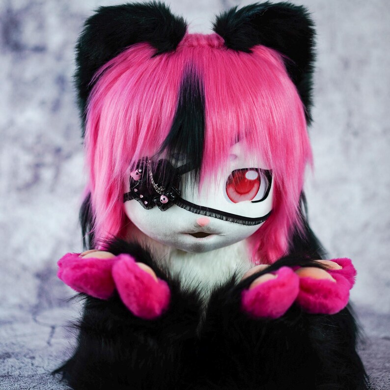 May include: A plush toy with black and pink fur, wearing a pink wig with a black streak. The toy has a white face with a black eye patch and one red eye. The toy is sitting with its paws in front of it.