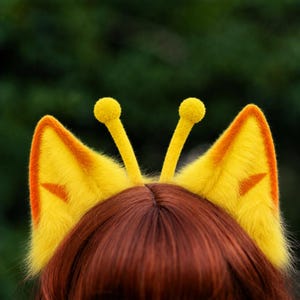 以下が含まれることがあります： 鮮やかな黄色のふわふわした尖った耳の内側にオレンジ色のアクセントがあり、先端に小さな球体がついた細長い黄色の触角が2本ついたヘッドバンド。ぼやけた緑色の背景を背に、ストレートのダークブラウンの髪の上に装着されている。
