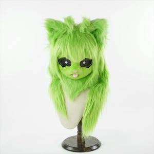 May include: A lime green furry cat mask with large black eyes, a pink tongue, and long, shaggy fur. The mask is displayed on a mannequin head with a wooden stand, set against a white background. The mask has a whimsical, cartoonish design.