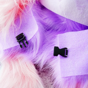 May include: Close-up of a fluffy, multi-colored fabric with pink, purple, and white hues. Two black plastic buckles are attached to the fabric, one with a three-pronged design and the other with a single-prong design. The fabric appears soft and textured.