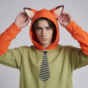 A person wearing an orange and green fox cosplay pullover hoodie with perked ears, posing playfully at a convention.