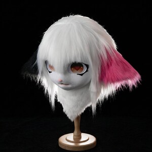 May include: A white furry mask with large, expressive eyes and a small pink nose. The ears are accented with black and pink fur. The mask is mounted on a wooden stand against a black background.