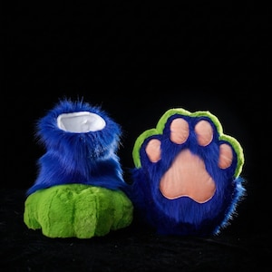 May include: A pair of furry blue and green paw-shaped costume feet. The feet have a blue upper portion with a white interior and a green sole. The paw pads are pink, and the outline is green.
