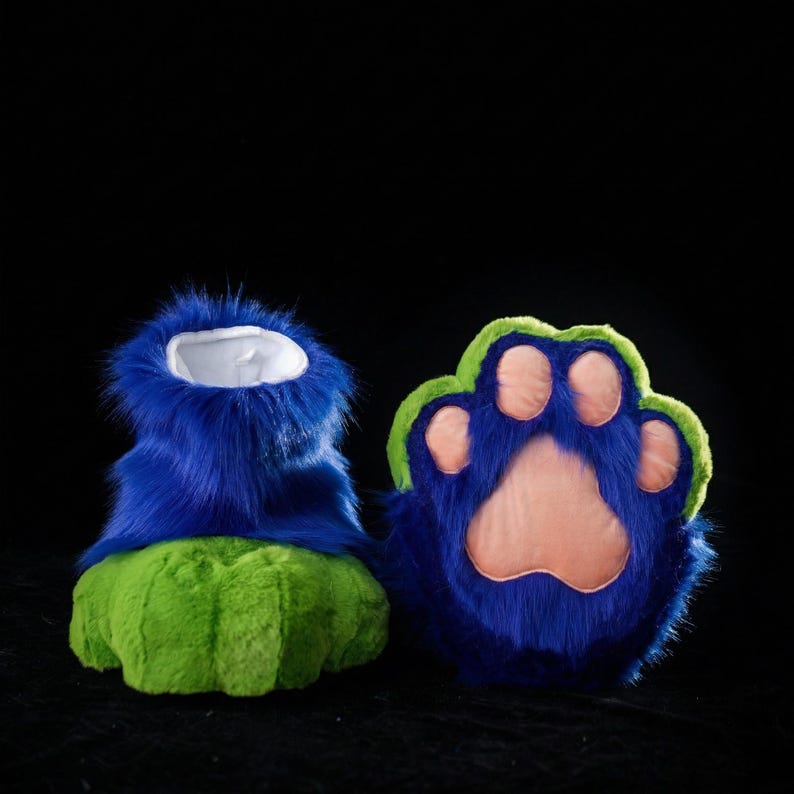May include: A pair of furry blue and green paw-shaped costume feet. The feet have a blue upper portion with a white interior and a green sole. The paw pads are pink, and the outline is green.