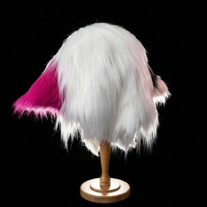 May include: A fluffy, white faux fur hat with a vibrant pink accent on one side. The hat is displayed on a wooden stand against a black background, showcasing its texture and shape. The hat's design is reminiscent of a playful, whimsical style.