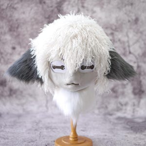 May include: A whimsical, handmade mask with a stylized face. The mask features a white, fluffy, fur-like hair, gray ears, and a white fur collar. The eyes are represented by rectangular shapes. The mask is mounted on a wooden stand.