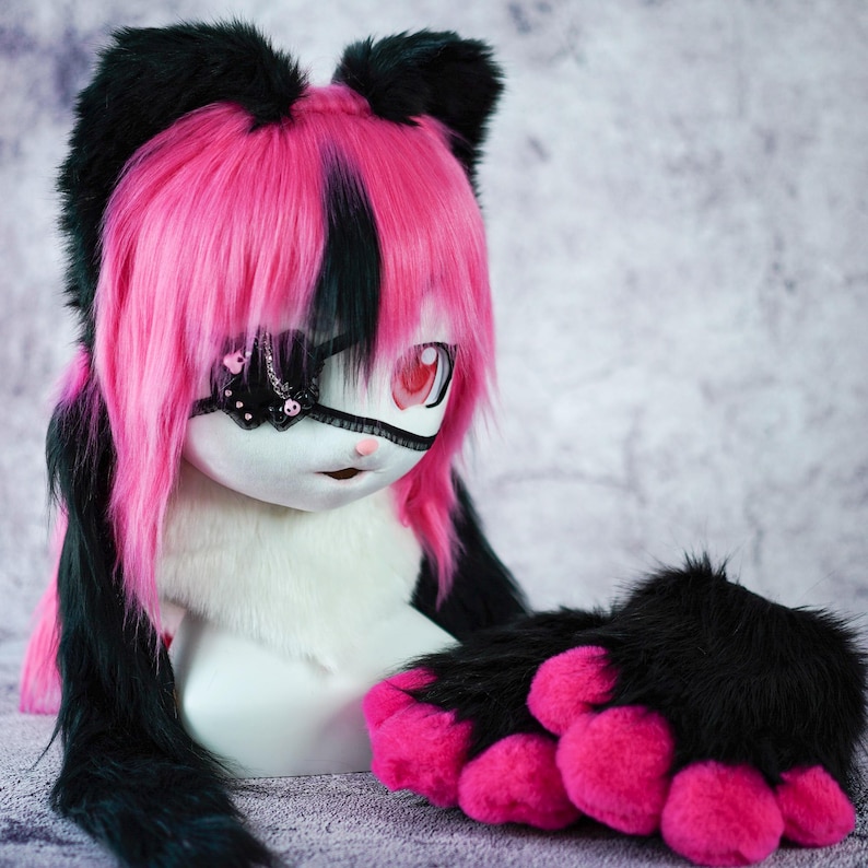 May include: A plush toy with black and pink fur, wearing a black eye patch with sparkly accents. The toy has pink paws and a pink and black wig.
