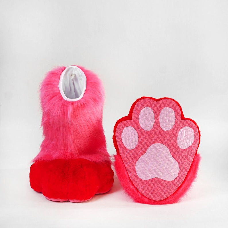 May include: A pair of plush slippers featuring bright pink faux fur on the upper section and a vibrant red base. The sole of the slipper is textured with a light pink paw print pattern, including four small toe pads and one large central pad.
