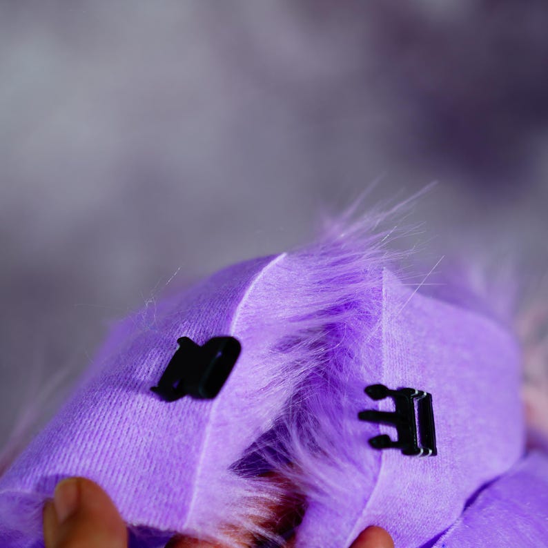 May include: Close-up of a purple fabric with black fasteners. The fabric has a soft, furry texture and is folded over, revealing the inside. The background is a blurred, light purple.