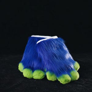 May include: A pair of furry blue and green costume feet. The feet are a vibrant blue with green toes and a white accent at the top. The plush material suggests a playful or character-based design.