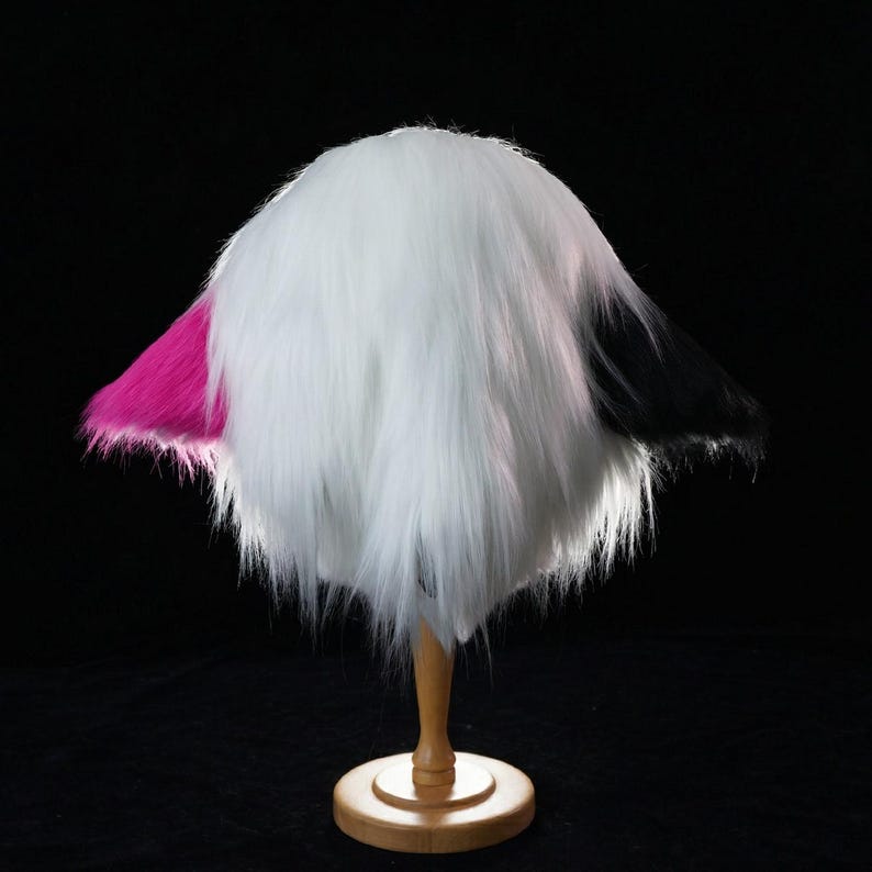 May include: A fluffy, faux fur hat in white, black, and hot pink. The hat is displayed on a wooden stand against a black background. The hat's design features a mix of colors and textures.