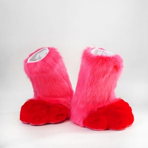 May include: A pair of ankle-high boots covered in long, vibrant pink faux fur. The boots feature rounded, solid red toe sections and a white interior lining visible at the top opening. The design is bulky with a thick, plush texture throughout.