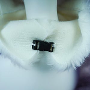 May include: A white faux fur collar featuring a smooth, cream-colored fabric lining on the interior side. The collar is secured at the front center with a small, black plastic side-release buckle, which is attached to the fabric with white thread.