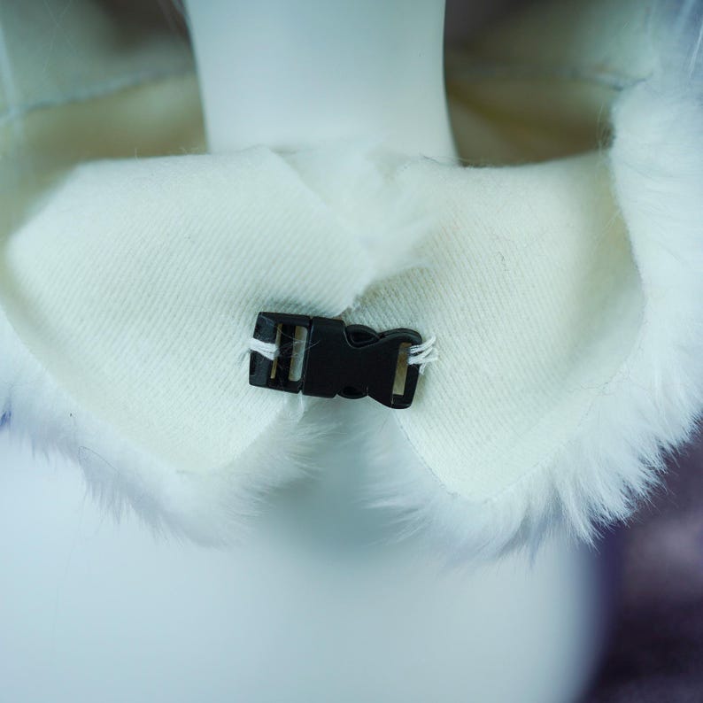 May include: A white faux fur collar featuring a smooth, cream-colored fabric lining on the interior side. The collar is secured at the front center with a small, black plastic side-release buckle, which is attached to the fabric with white thread.