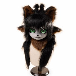 May include: A creature mask featuring long black faux fur, brown inner ear accents, and a brown neck ruff. The face is light grey with a small black nose and large, expressive green eyes with intricate iris detailing, set on a dark wooden stand.