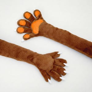May include: Two brown plush paw gloves with orange paw pads and white claws are displayed against a white background. The gloves are designed to resemble animal paws, with detailed stitching and a soft, furry texture. The gloves are likely part of a costume or novelty item.