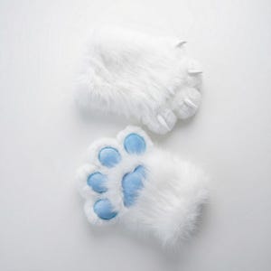 Op de afbeelding: Een paar witte nepbont poothandschoenen met lichtblauwe voetzolen en witte klauwen. De handschoenen zijn ontworpen om op dierenpoten te lijken, met een zachte textuur en een speelse uitstraling.