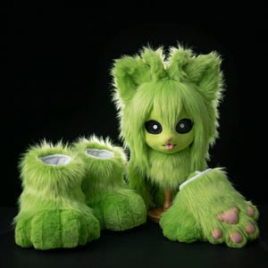 May include: A green furry doll head with long hair, large black eyes, and a pink tongue, along with matching green furry feet and leg coverings. The doll head is on a wooden stand. The background is black.