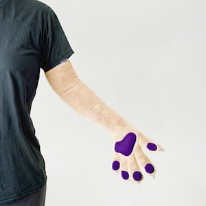 Peut inclure: Une manche de bras en fourrure beige avec un coussinet de patte violet et des haricots d'orteils violets. La patte a des griffes blanches. La manche est portée sur le bras d'une personne portant un t-shirt noir.