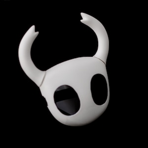 May include: A white mask with two large, oval eye holes and two curved, horn-like protrusions. The mask is made of a smooth, matte material and is set against a black background. The mask's design is simple and stylized.