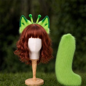 May include: A set consisting of a headband with bright green faux fur animal ears and two antennae, worn on a mannequin head with wavy auburn hair, accompanied by a matching long, fluffy, bright green faux fur tail accessory.
