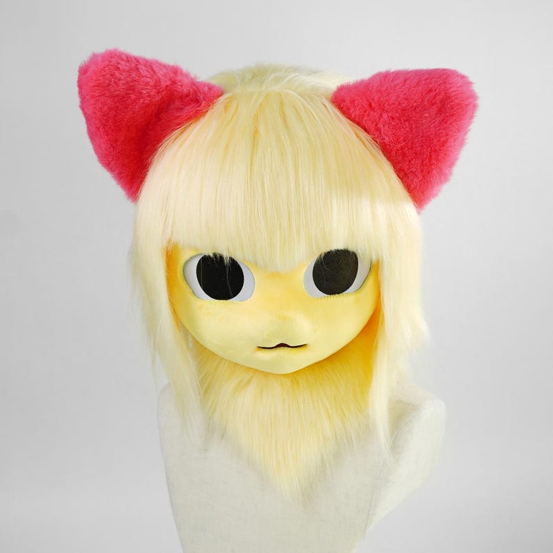 May include: A full-head mask featuring pale yellow synthetic fur and hair with straight bangs. The mask has large, round, black eyes and a small mouth. Two bright pink, fuzzy, triangular ears are attached to the top of the head.