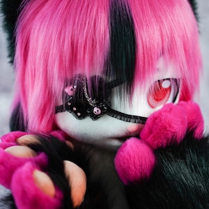 May include: A pink and black furry creature with a black eye patch and pink paws. The creature has a pink and black striped hairstyle.