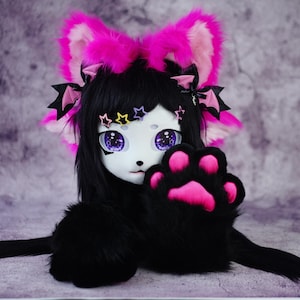 May include: A black and white fursuit head with large purple eyes, black hair, and pink accents. The fursuit has a paw with pink pads and pink and black bat-shaped hair accessories. The ears are pink and white.