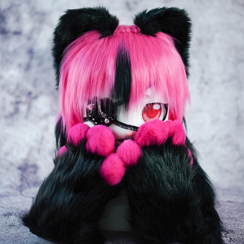 May include: A plush toy with black fur and pink ears, wearing a pink and black wig with a black eye patch. The toy has a white face with red eyes and is holding its paws in front of its face.