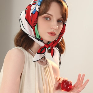May include: A woman with brown hair is wearing a white, red, and blue floral headscarf. She is holding a red heart-shaped object in her hands.