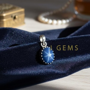 Op de afbeelding: Een zilveren hanger met een blauwe ster saffier edelsteen. De edelsteen heeft een zesstralig sterpatroon. De hanger ligt op een donkerblauwe fluwelen stof. Het woord "GEMS" is zichtbaar.