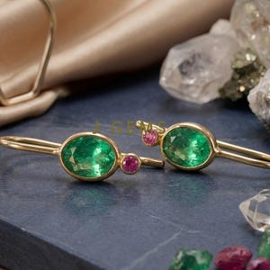 Puede incluir: Pendientes de oro con esmeraldas ovaladas y detalles de rubíes. Los pendientes tienen un diseño delgado y alargado. Esmeraldas y rubíes sueltos dispersos cerca. Los pendientes están sobre una superficie azul oscuro.