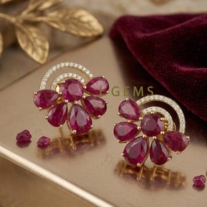 May include: Gold-coloured stud earrings with a cluster of deep red, teardrop-shaped gemstones. Each earring has a halo of small, clear stones around a curved design. Small loose gemstones are scattered around the earrings.