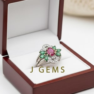 Può includere: Un anello d'argento con un rubino ovale centrale, circondato da smeraldi verdi e piccoli diamanti. L'anello è esposto in un portagioie in legno scuro aperto. Il testo "J GEMS" è visibile.