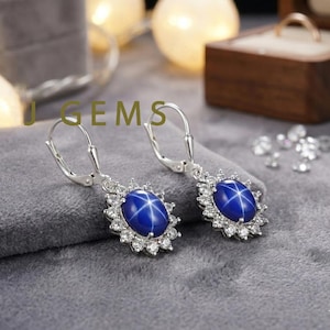 May include: A pair of silver leverback earrings. Each earring features a large, oval, deep blue gemstone with a star-like pattern, surrounded by a halo of small, clear gemstones. The earrings are displayed on a gray surface.