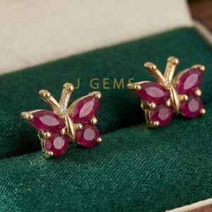 May include: A pair of gold-colored butterfly stud earrings. Each earring features a butterfly design with ruby-colored gemstones and a small diamond accent. The earrings are displayed on a dark green surface.
