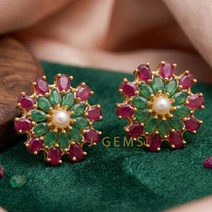 Op de afbeelding: Een paar goudkleurige oorstekers. Elke oorbel heeft een bloemenontwerp met afwisselende robijn- en smaragdgroene edelstenen rond een centrale parel. De oorbellen zijn geplaatst tegen een donkergroene achtergrond.