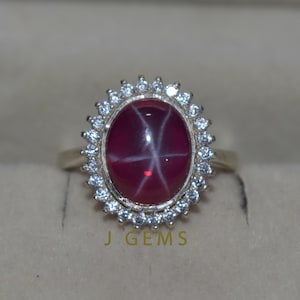 Peut inclure: Une bague en or ornée d'une grosse pierre précieuse ovale rouge foncé avec un motif en étoile. La pierre précieuse est entourée d'un halo de petites pierres précieuses claires. Le texte "J GEMS" est visible en bas de l'image.