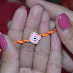 May include: A close-up of a bracelet featuring a pink flower-shaped charm with a central eye design. The charm is adorned with small, sparkling stones and set on a twisted orange and yellow cord. The bracelet is held between two hands.