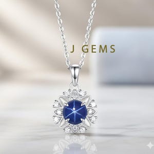 Op de afbeelding: Een zilveren ketting met een hanger met een donkerblauwe ster saffier edelsteen. De saffier is omgeven door een halo van kleine, heldere edelstenen en gezet in een bloemvormig ontwerp. De tekst "J GEMS" is zichtbaar.