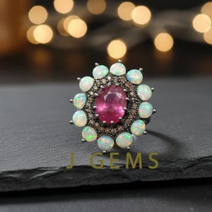 Peut inclure: Une bague en argent ornée d'une pierre rose ovale au centre, entourée d'un halo d'opales ovales plus petites. Les opales ont une base blanche laiteuse avec des éclats de couleur. La bague est posée sur un fond sombre avec le texte "GEMS".