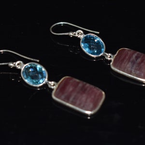 May include: A pair of silver dangle earrings. Each earring features a blue oval gemstone and a rectangular, maroon gemstone. The earrings are displayed against a black background.