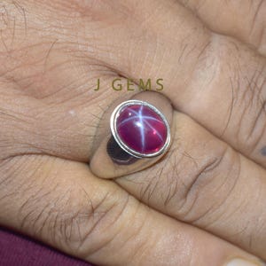 May include: A silver ring featuring a large, oval, deep red gemstone with a six-rayed star pattern. The ring is set in a simple, polished silver band. The text "J GEMS" is visible in the image.