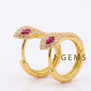 May include: Gold-colored hoop earrings with a snake design. The snake's head is adorned with small, clear stones and a central, marquise-cut, ruby-colored stone. The earrings are set against a white background, with the text "J GEMS" visible.