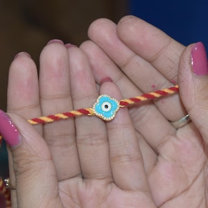 May include: A gold-colored bracelet with a blue eye-shaped charm and a red and yellow twisted cord. The charm has a white center and is surrounded by a light blue border. The cord is held by a person.