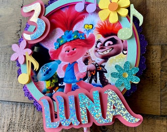 May include: A pink and purple cake topper with a picture of the Trolls characters, a musical note, and the name "LUNA" in glittery pink letters. The number "3" is also on the topper.