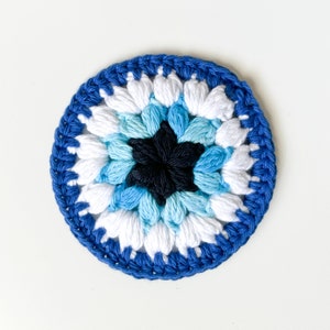 May include: A crocheted blue and white eye charm with a black star in the center. The charm is round and has a textured surface.