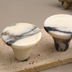 May include: Two round cabinet knobs made of polished white marble with prominent dark grey and black veining. Each knob features a smooth, circular top surface and a tapered base, showcasing the natural stone patterns and glossy finish.
